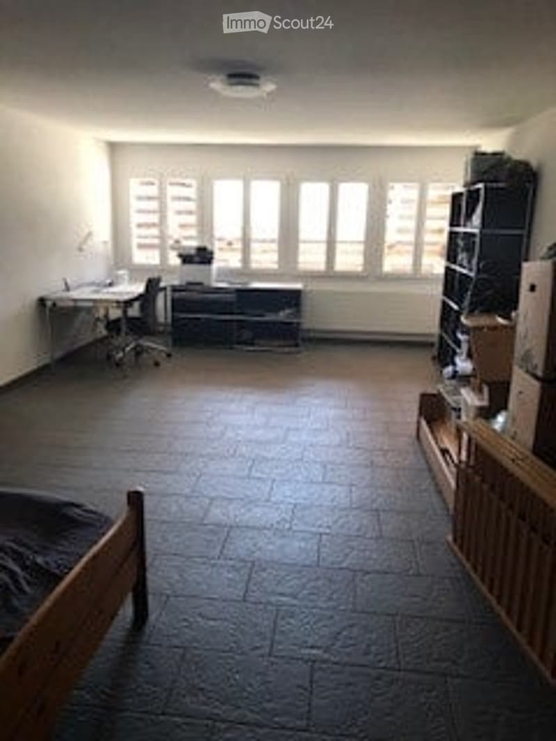 A spacious, well-lit room with large windows, a wooden bed, a desk with a chair, a cabinet, and a shelf. The floor is tiled, and the walls are white.