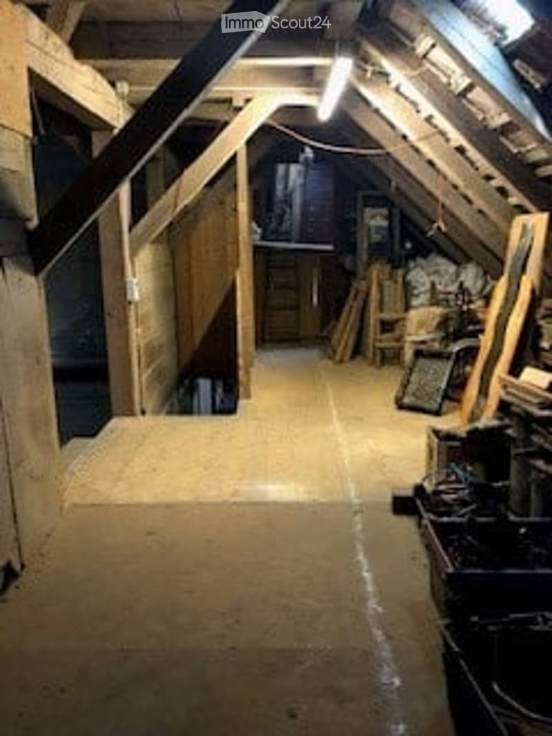 An attic space with exposed wooden beams, a staircase leading up, and a few scattered items like a chair and a small table.