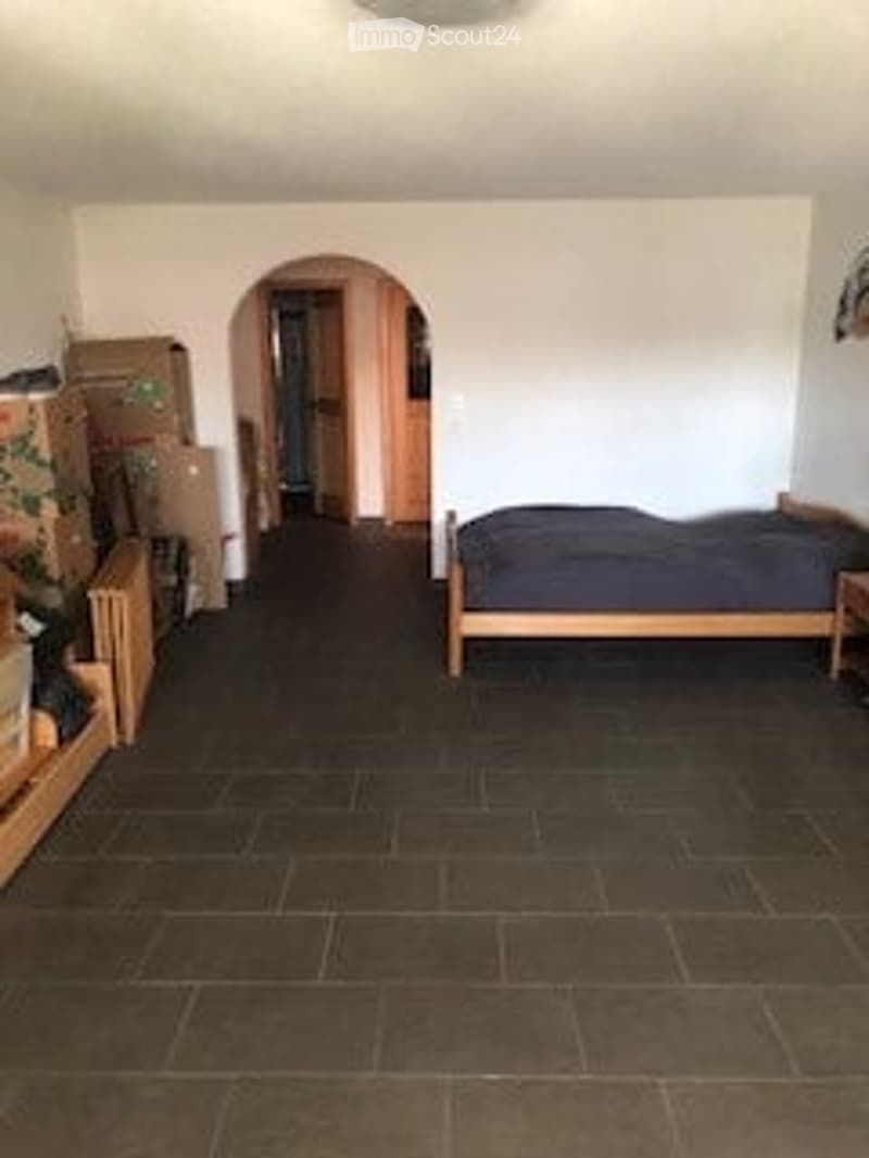 A furnished bedroom with a bed, nightstand, and wardrobe. The room features dark tiled flooring and white walls.