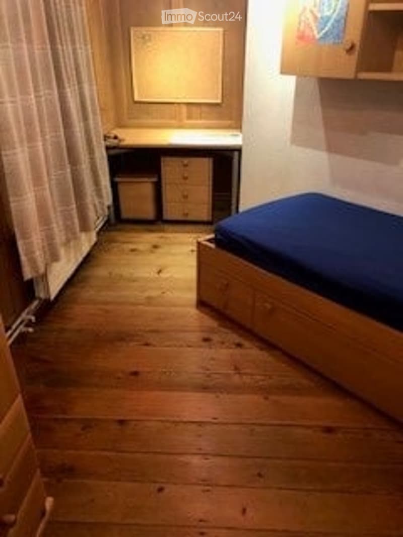 A small, furnished room with wooden flooring, a single bed with blue bedding, a desk with a chair, and a small cabinet.