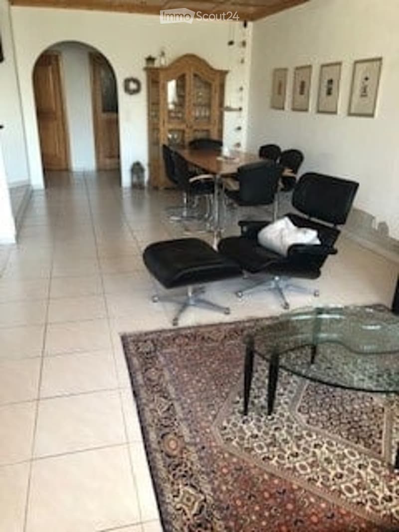 Bright living room with white walls, tiled floor, and large windows. Features a black leather sofa, a glass coffee table, a dining table with chairs, and a wooden cabinet with glass doors.