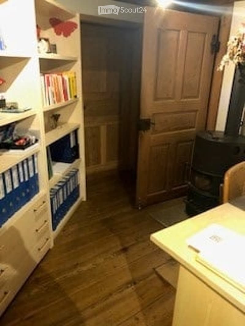 A cozy room with wooden floors, a large bookshelf filled with books, a wooden door, a small table with a chair, and a fireplace in the corner.
