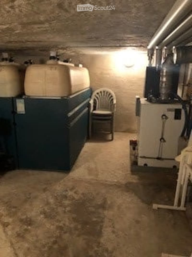 Utility room with water tanks, a boiler, and a single chair. Exposed pipes and a concrete floor.