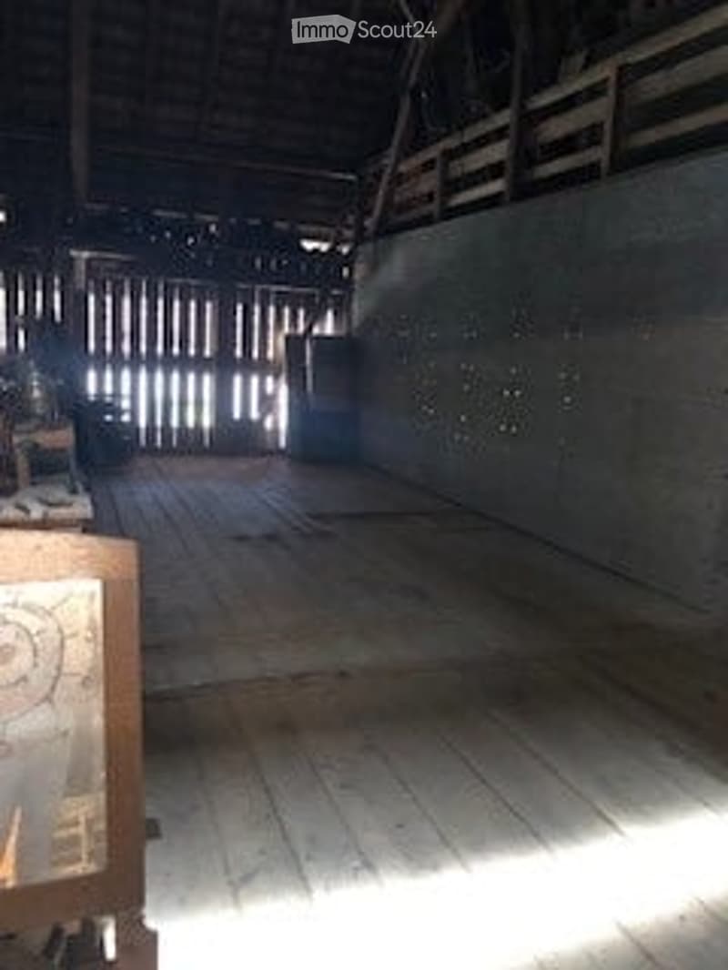 Empty room with wooden floor and exposed wooden beams on the ceiling. Natural light streams in from a large window or opening at the far end.