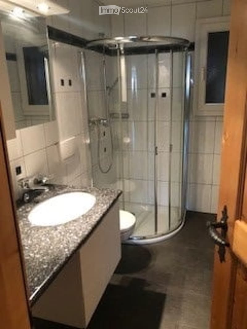 Modern bathroom with a white tiled shower stall, a sink with a chrome faucet, and a toilet. The walls are tiled in white, and the floor is dark.