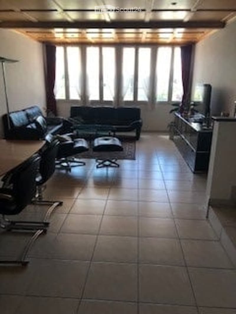 Modern living room with large windows, tiled floor, black sofa, coffee table, chairs, ottoman, floor lamp, and TV stand.