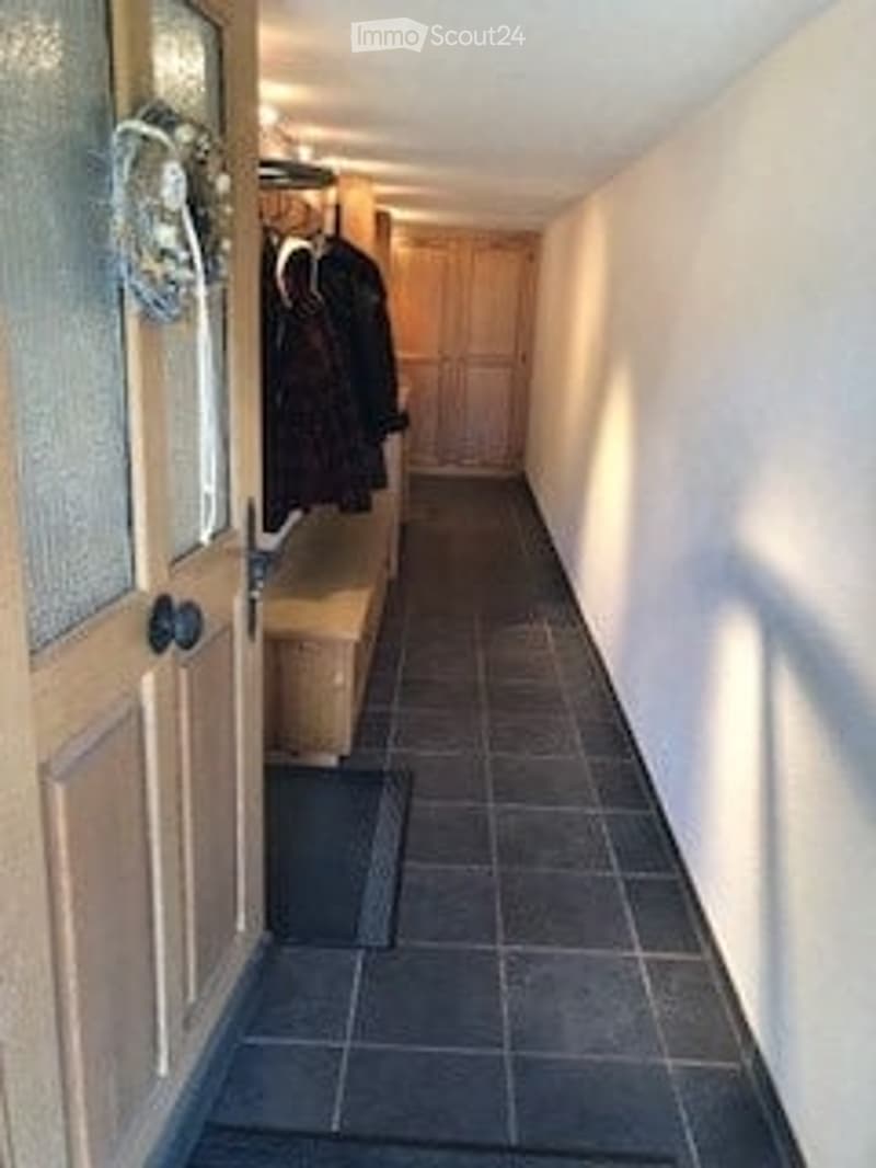 A narrow hallway with tiled flooring, light-colored walls, and a coat rack with jackets hanging on it. The hallway leads to a door at the end.