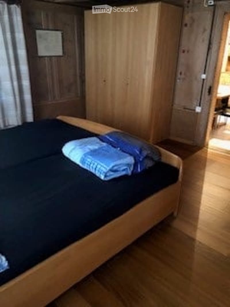 Cozy bedroom with wooden bed frame, dark bedding, and a large wooden wardrobe. Wooden flooring and warm lighting.