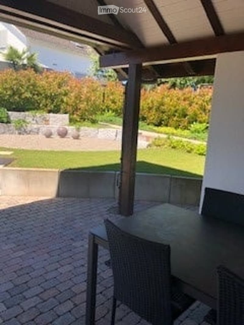 A covered outdoor patio with a brick floor, a round wooden table, and black wicker chairs. The background shows a well-maintained garden with green grass, shrubs, and a stone wall.