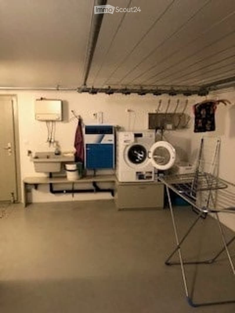 Utility room with a washing machine, dryer, sink, and a small stool. The room has exposed pipes and a simple, functional design.