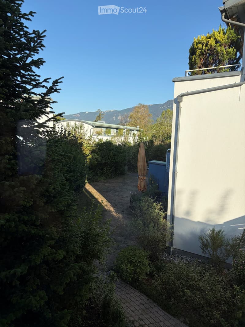 A narrow garden pathway lined with greenery and shrubs, leading to a building with a white exterior and a balcony. The area is surrounded by trees and has a clear blue sky and distant mountains in the background.