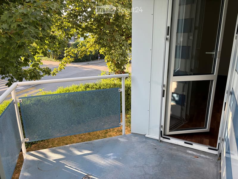 Balcony with metal railing, open sliding glass door leading inside, concrete floor, greenery visible in background.