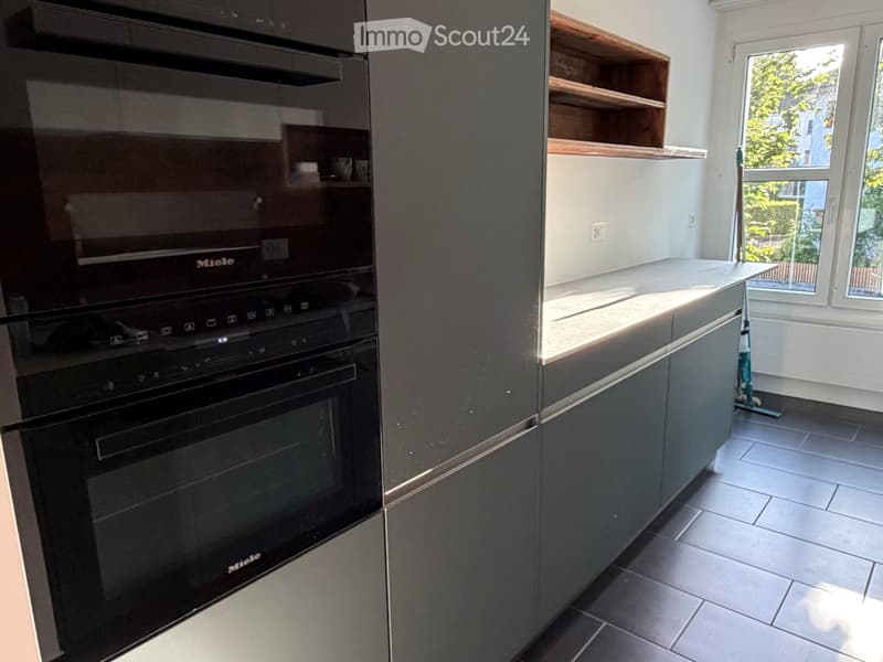 Modern kitchen with built-in oven, grey cabinets, and wooden shelves. Large windows provide natural light.