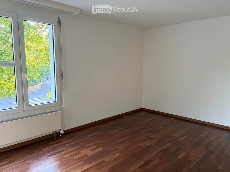 Empty room with wooden flooring, white walls, and large windows showing greenery outside.