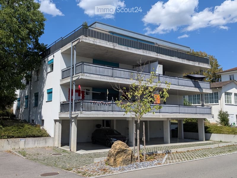 Modern three-story apartment building with balconies, garage, and surrounding greenery.