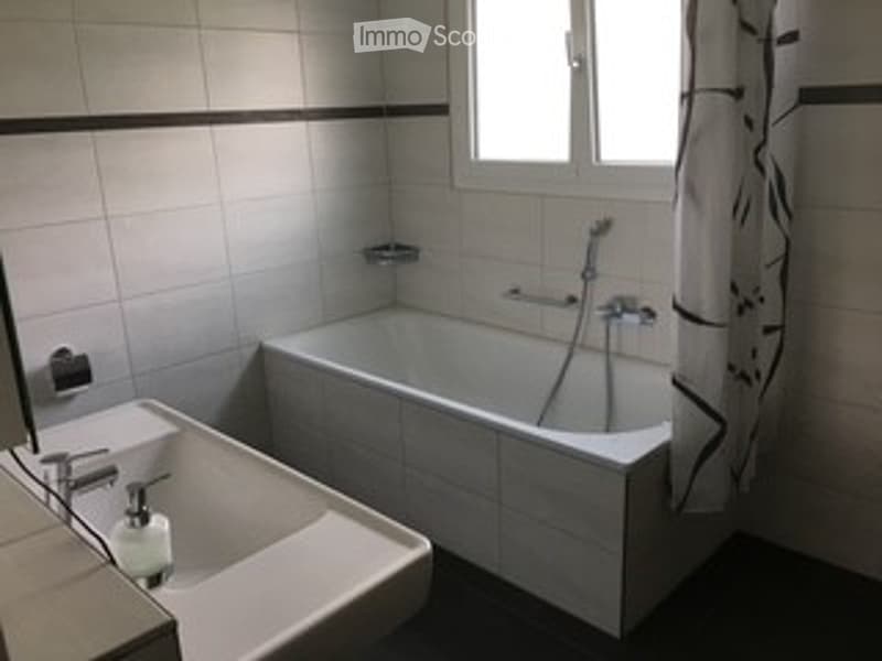 A clean, modern bathroom with a bathtub, toilet, and a window with a curtain. The walls are tiled in white with black trim, and the floor is dark.