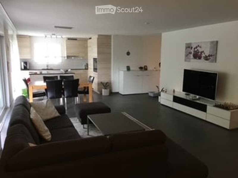 Modern open-plan living and dining area with a dark grey sofa, coffee table, ottoman, flat-screen TV on a white stand, and a kitchen island with dining chairs.
