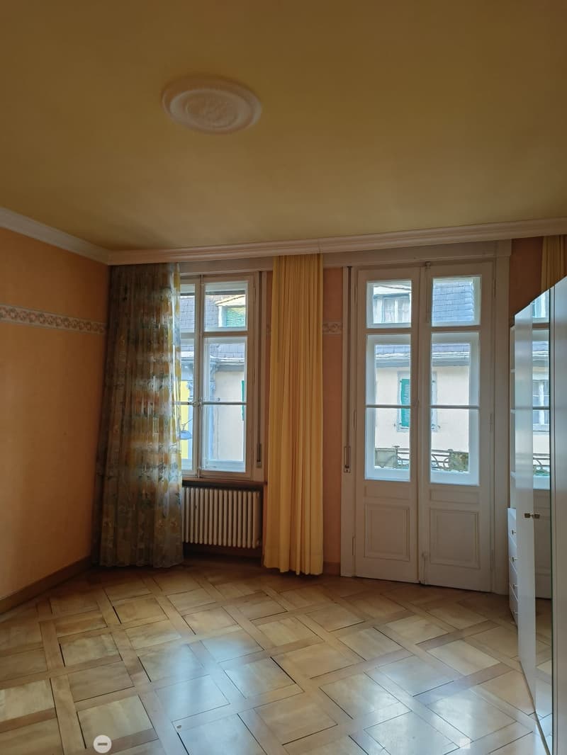 Empty room with wooden tiled floor, large windows with curtains, radiator, and double doors leading to a balcony.