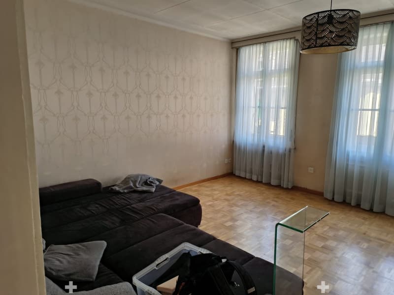 Living room with a dark grey sectional sofa, a glass coffee table, and a patterned wall design. The room features wooden flooring and large windows with sheer curtains.
