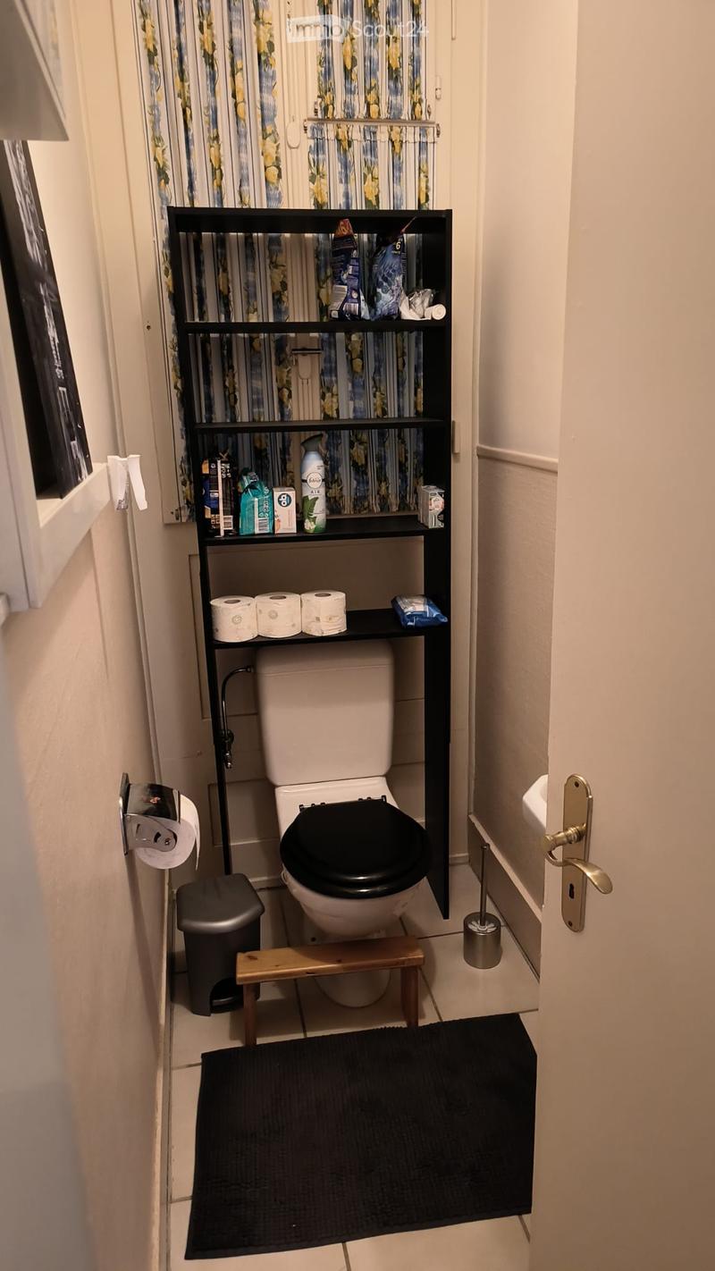 Compact bathroom with a toilet, black shelf holding toiletries, toilet paper, a small wooden stool, a trash can, and a black bathroom rug.