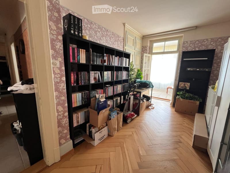 A room with wooden flooring, a large bookshelf filled with books, a small table with a plant, a vacuum cleaner, and a refrigerator. The room has a door leading to a balcony or outdoor area.