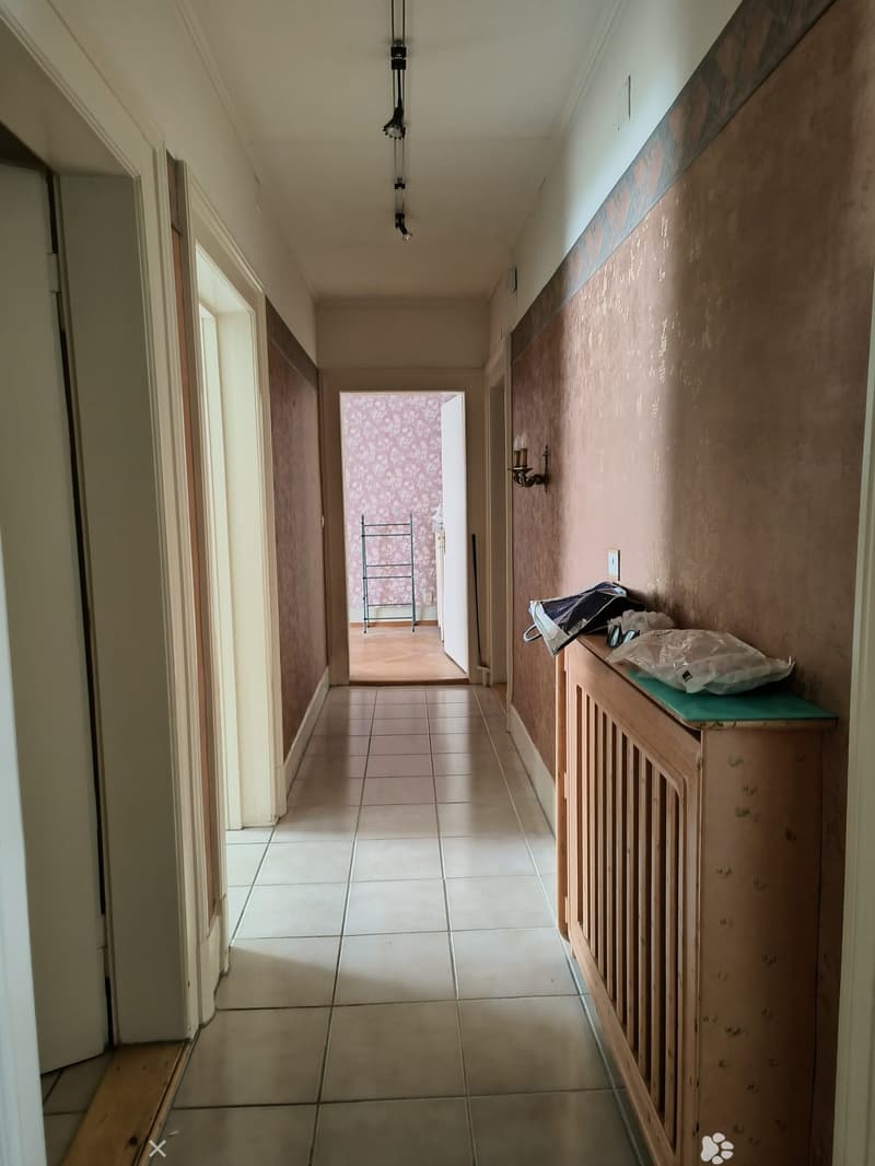 Empty hallway with tiled floor, beige walls, and a radiator on the right side. A small green shelf holds a few items, and a metal drying rack is visible in the background.