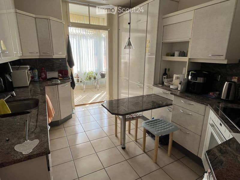 Modern kitchen with white cabinets, granite countertops, and stainless steel appliances. Features a central island with a black granite top, two stools, and a pendant light. The kitchen has a large window with blinds and a view of a tiled patio area.