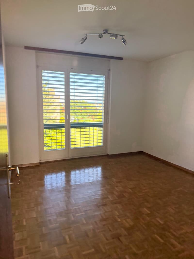 Empty room with wooden flooring, white walls, and large windows with blinds. The room has a modern ceiling light fixture and a door leading to an outdoor area.