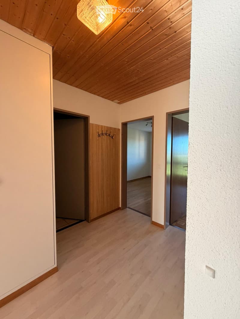 Empty hallway with wooden flooring, white walls, and several doors leading to other rooms. The ceiling has wooden paneling and a hanging light fixture.
