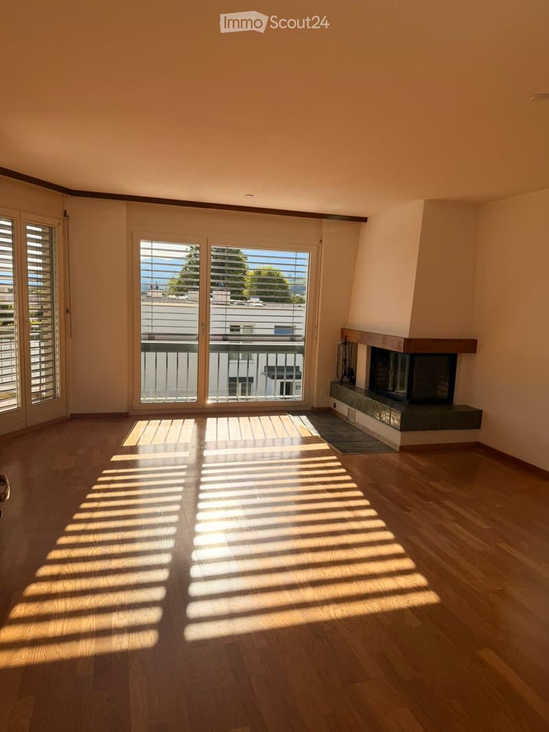 Empty living room with wooden flooring, large windows, and a fireplace. Sunlight streams in through the windows, casting shadows on the floor. The room features a balcony accessible through glass doors.
