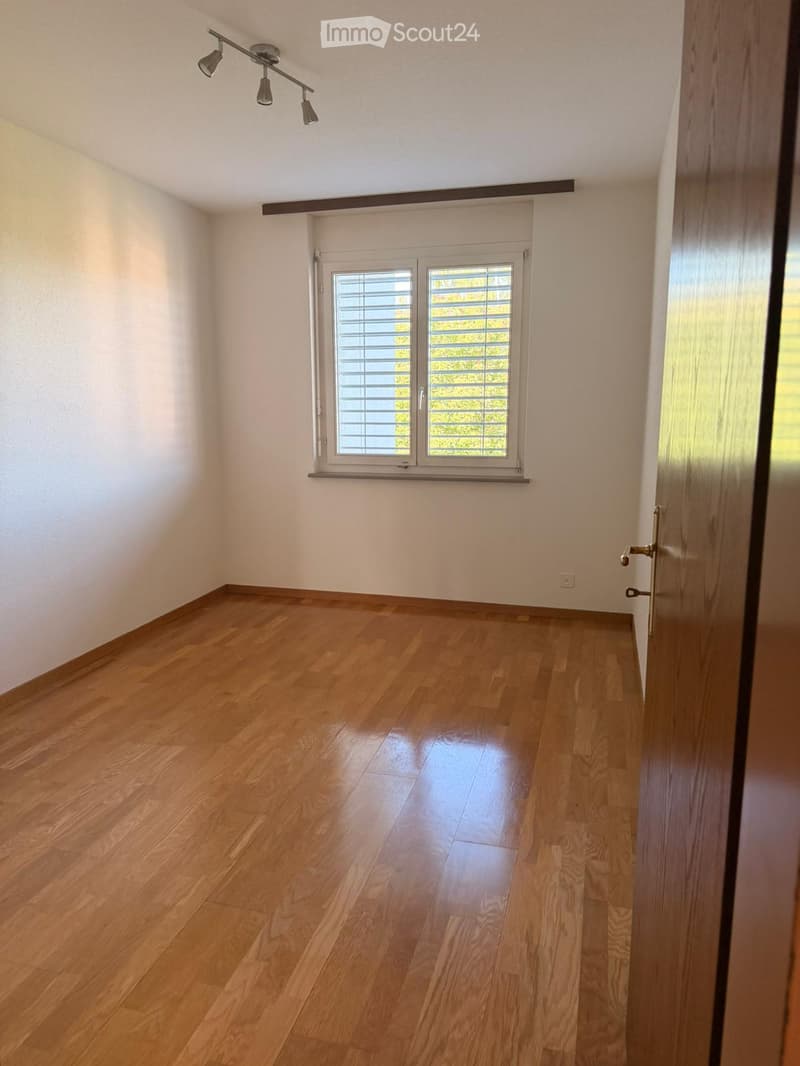 Empty room with light wooden flooring, white walls, and a large window with blinds. The room features a door on the right side and a track light fixture on the ceiling.