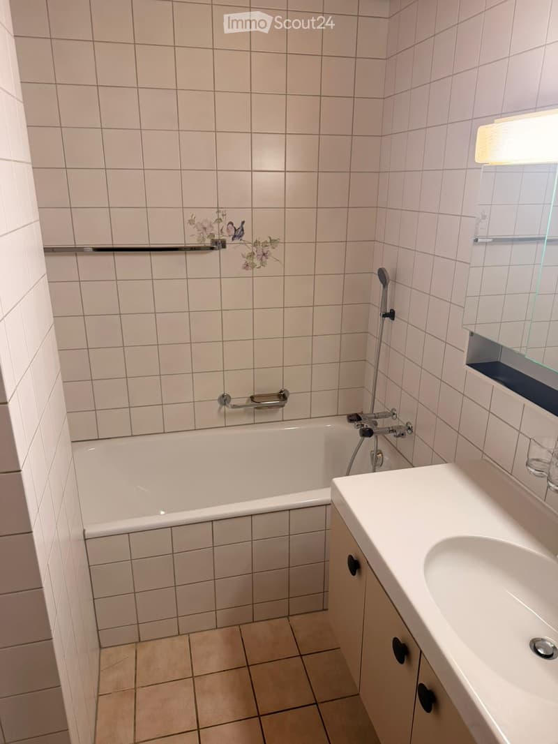 Bathroom with white tiled walls and floor, featuring a bathtub with a handheld shower, a sink with a cabinet, and a window with a frosted glass pane.