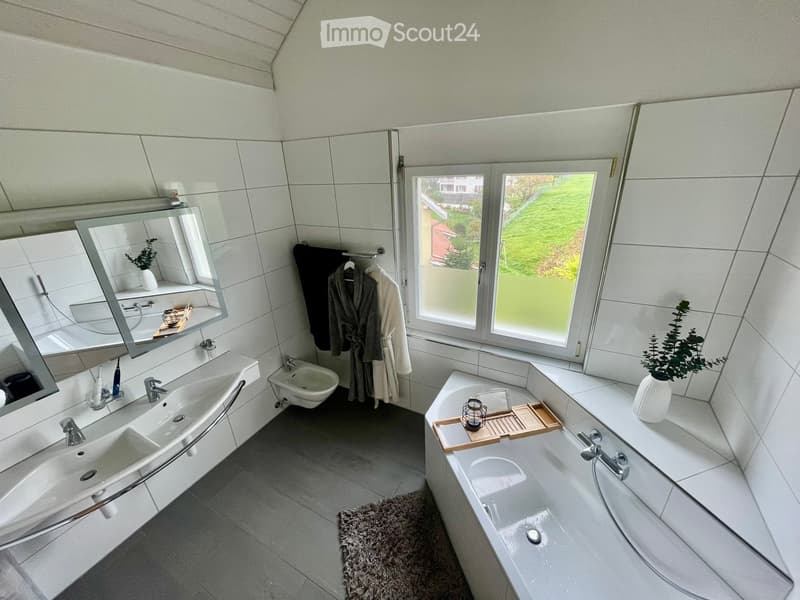 Modern bathroom with a large window, double sink, bathtub, and a mirror. Includes a small plant, a towel rack with robes, and a decorative tray on the bathtub.