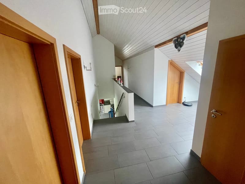 A modern hallway with grey tiled flooring, white walls, wooden doors, and a small shelf with decorative items visible in the background.