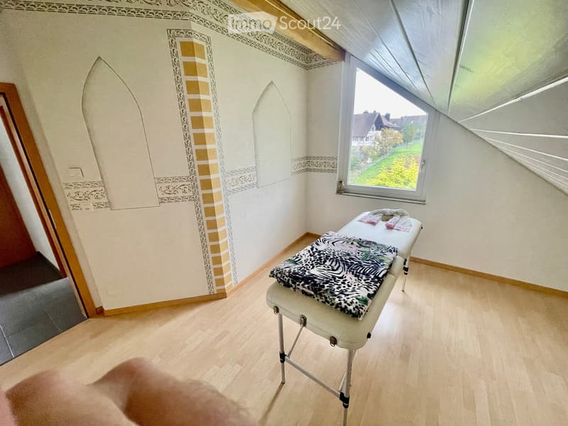 A small, well-lit room with wooden flooring, a massage table covered with a patterned cloth, and a few towels placed on it. The room features decorative wall panels and a large window offering a view of the garden.