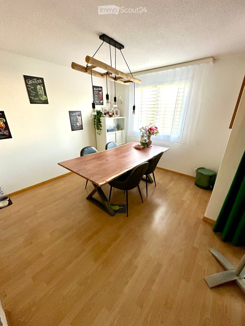 Dining room with a wooden dining table, four chairs, pendant light fixture, white cabinet, vase with flowers, and a green stool near the window.