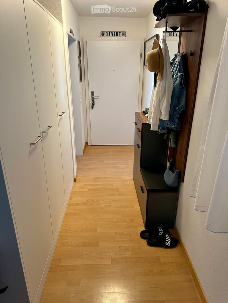 A clean, modern hallway with light wooden flooring, white walls, and built-in storage. A coat rack holds jackets and hats, and a small bench with drawers is visible.