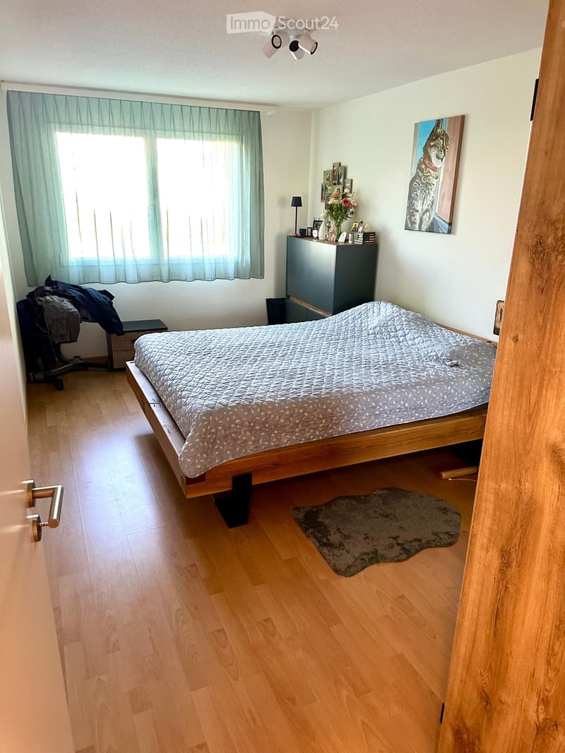Cozy bedroom with wooden flooring, a large bed with a patterned duvet, a nightstand with a lamp, a picture frame on the wall, and a small rug on the floor.