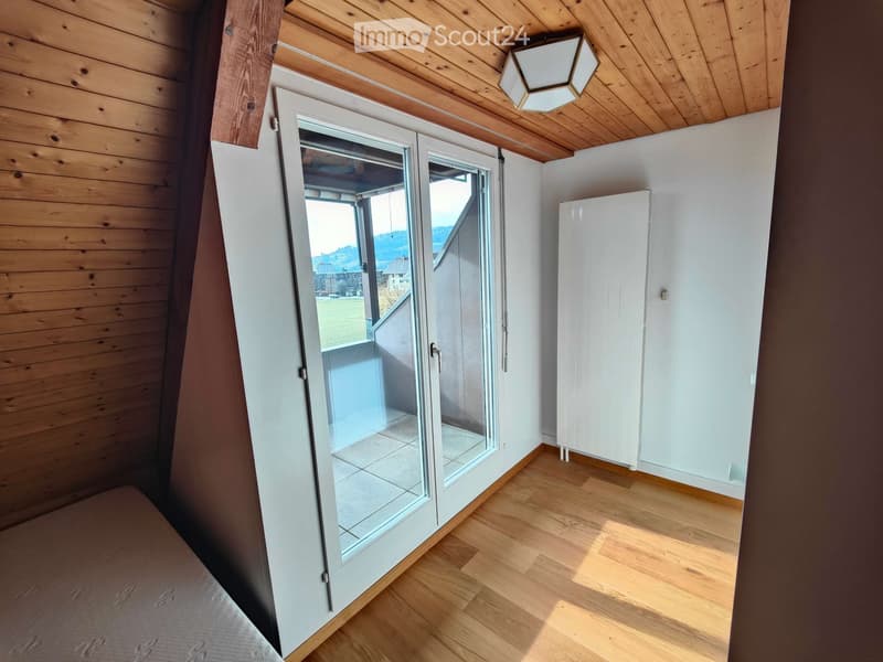 Bright bedroom with wooden floors, slanted wooden ceiling, large sliding glass doors leading to a balcony with a scenic view, and a white wardrobe.