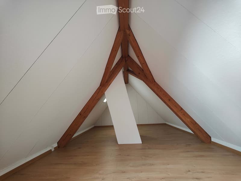 Empty attic space with wooden beams and slanted white walls, featuring a wooden floor and exposed wooden structural beams.