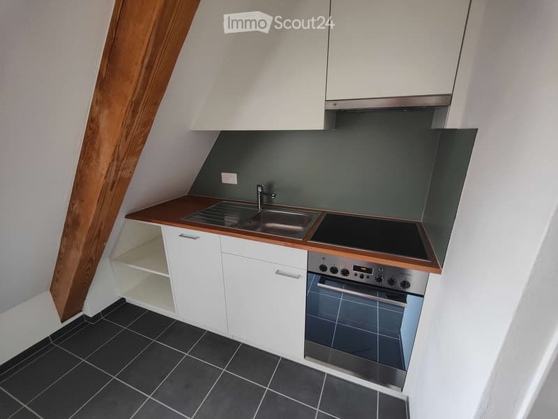 Compact kitchen with white cabinets, dark countertops, a sink, and an oven, featuring a slanted ceiling and black tiled floor.