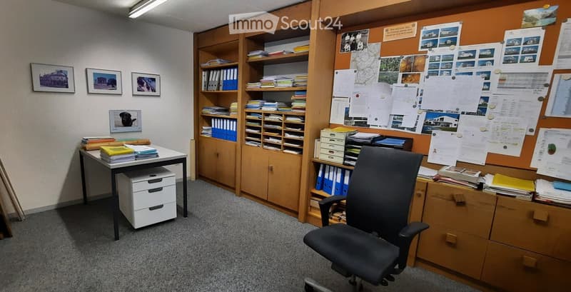 Empty office room with white wall, cabinets, bookshelf, picture frames, office chair, desk, drawers, ceiling lamp, posters on the wall
