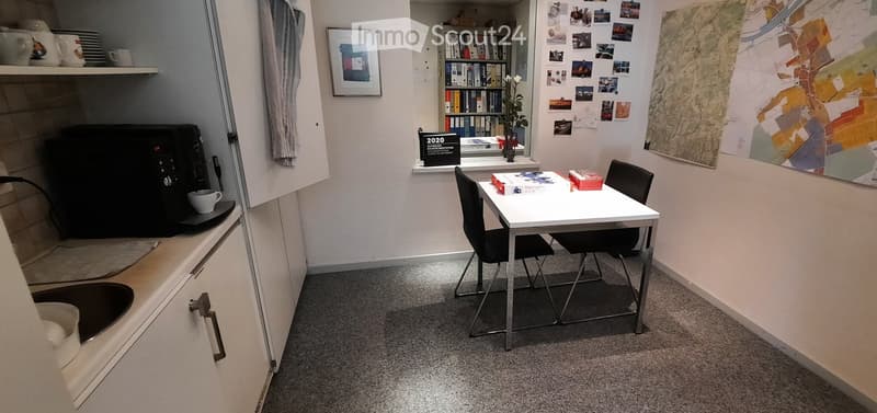 Small room with table, chairs, sink, coffee machine, bookshelves, artwork, small window with white curtain