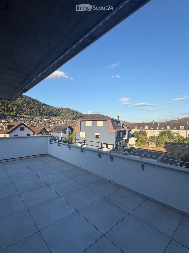 Modern rooftop terrace with tiled flooring, white walls, and a metal railing, overlooking a town with houses and hills in the background.