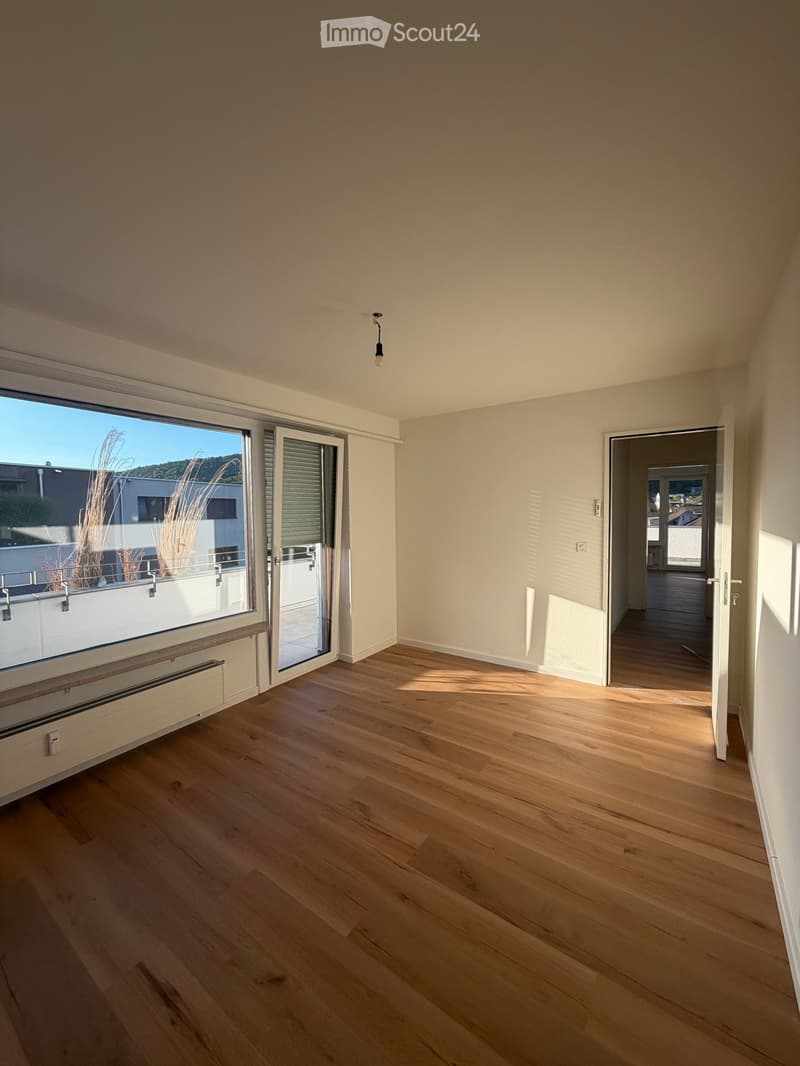 Empty room with wooden flooring, large window with view, sliding glass door leading to balcony, and a radiator under the window.