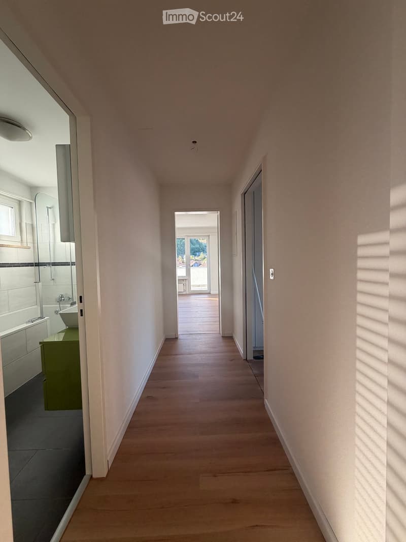 A narrow hallway with light wooden flooring and white walls, leading to a room with large windows and a view of greenery. A bathroom is visible on the left side with a bathtub, sink, and green cabinet.