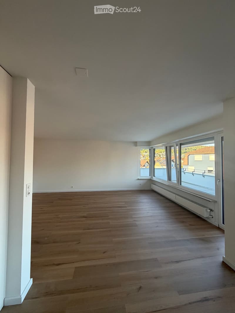 Empty room with wooden flooring, white walls, and large windows offering a view of the outside area.