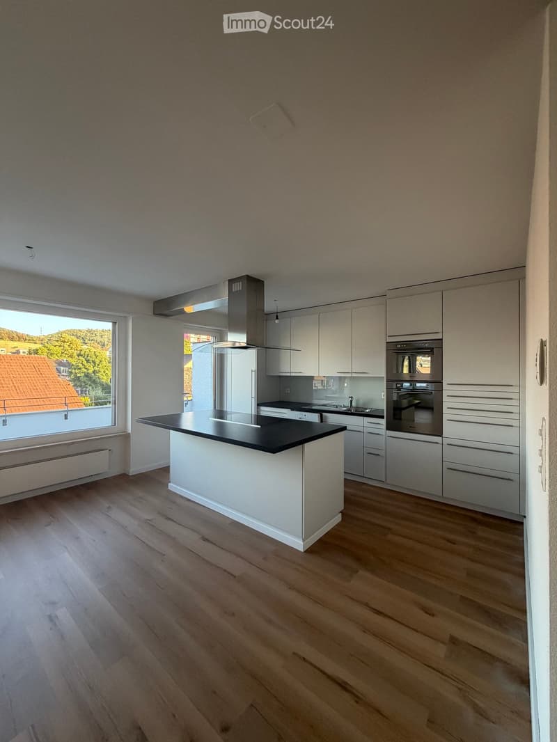 Modern kitchen with light-colored cabinets, a central island, and stainless steel appliances including an oven and refrigerator. The space features wooden flooring and large windows offering a view of the outdoors.