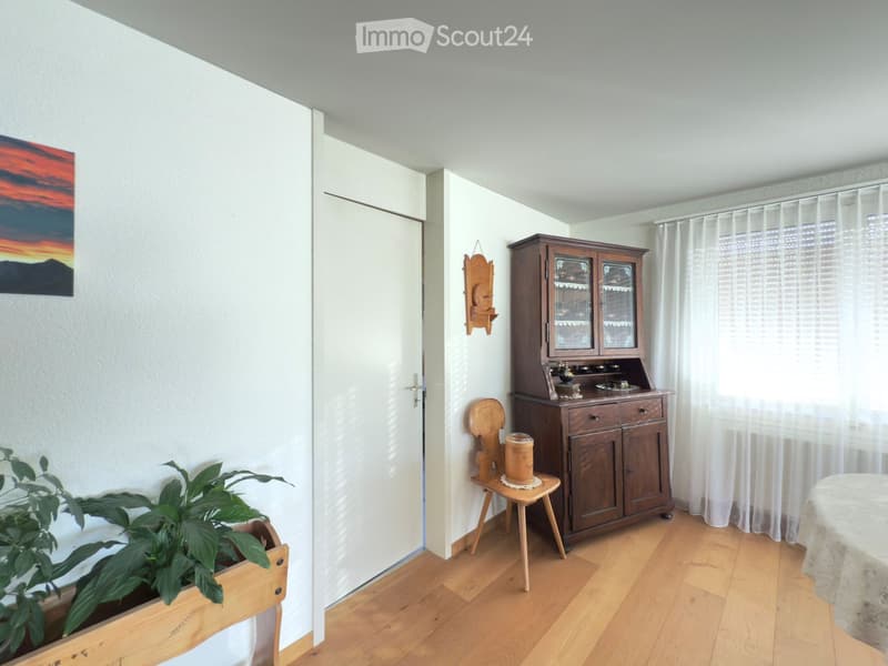 A well-lit room with wooden flooring, a large wooden cabinet with glass doors, a small wooden stool, a table with a tablecloth, a potted plant, and a painting on the wall.
