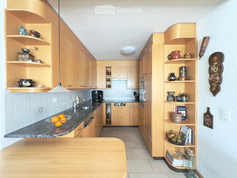 The kitchen features light wooden cabinets, a granite countertop, and a sink with a modern faucet. Shelves display decorative items and books, and a bowl of oranges sits on the counter.